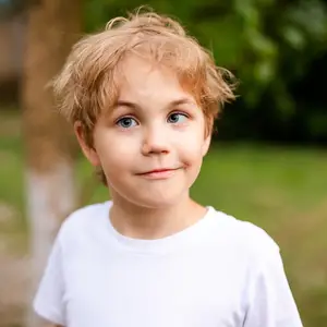 A smiling boy with blonde hair and blue eyes wearing a white shirt is standing in an outdoor area with grass and trees. The boy has a slight smile and a playful expression. The background is slightly blurry, with some trees and grass. The boy's hair is slightly messy, and the sunlight is reflecting on his face. The image has a warm and cheerful tone.