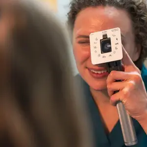 A woman optometrist holds an ophthalmoscope against her eye while examining another person's eye.