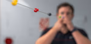 A man in a black shirt is holding a red and green object in his hands and seems to be playing with it while standing in front of a white wall with a red circle and a white board with some text on it.