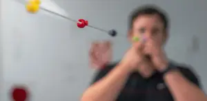 A man in a black shirt is holding a red and green object in his hands and seems to be playing with it while standing in front of a white wall with a red circle and a white board with some text on it.
