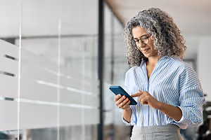 An adult woman with curly gray hair wearing glasses and a striped shirt is looking at her phone and smiling while standing in front of a glass wall in an office.