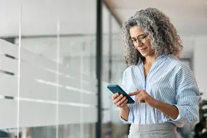 An adult woman with curly gray hair wearing glasses and a striped shirt is looking at her phone and smiling while standing in front of a glass wall in an office.