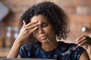 A woman with curly hair wearing a blue top with a floral design sitting at a desk with a laptop and holding a pair of glasses and looking at the camera with a sad face