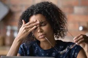 A woman with curly hair wearing a blue top with a floral design sitting at a desk with a laptop and holding a pair of glasses and looking at the camera with a sad face