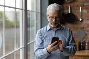 An older man is standing in the kitchen, holding his phone, and looking out of the window.