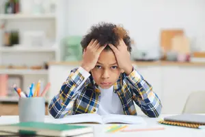 A boy sitting at a desk, holding his head in his hands and appearing to be in distress.