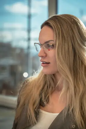 A woman wearing eyeglasses is standing in front of a glass window and looking at something in front of her.