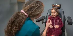 A young girl with curly hair and a pink headband sitting in a dental chair and a woman in blue scrubs holding a dental tool is smiling and looking at the girl