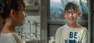 A young girl in a white shirt with the word BE and a cat print on it is smiling and looking at something outside a glass window with a display of glasses on a white shelf in the background.
