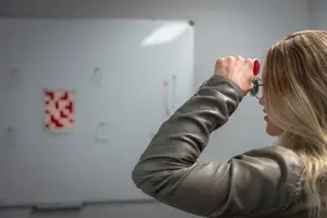 A woman wearing glasses with red lenses holds a pair of goggles up to her eyes while standing in front of a white board with several red squares.