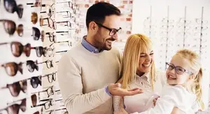 Three people, including a man, a woman, and a young girl, are standing together and smiling. They are probably inside a shop. The man is holding the young girl's hand, and the woman is holding the man's arm. Behind them is a white wall with a brick pattern and several pairs of eyeglasses hanging on it.