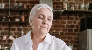 An older woman in a white shirt stands in front of a brick wall, with shelves of cups and bottles behind her, looking thoughtfully at something in the distance.