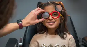 A young girl in a beige sweater is trying on red and black-framed glasses in an optometrist's office.