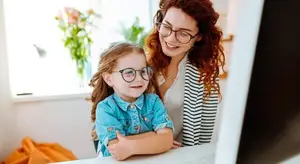 A woman and a girl are sitting on a chair in front of a monitor, smiling and looking at each other