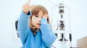 A young girl is holding her eyes while wearing a blue sweater in front of a white wall and a blurry background of an eye test machine