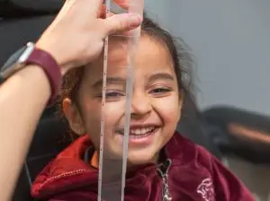 A young girl is having her vision checked by an adult wearing a watch and holding a clear plastic ruler near her eyes.