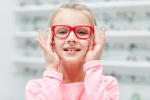 A young girl wearing red glasses and smiling at the camera while standing in a room with a blurry background of shelves with glasses