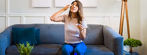 A woman sitting on a couch, holding a tissue, and seems to be applying something to her eye.