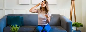 A woman sitting on a couch, holding a tissue, and seems to be applying something to her eye.
