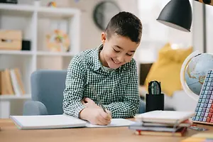 A young boy is sitting on a chair at his desk and smiling as he writes in a notebook.