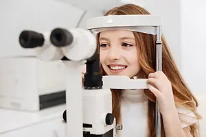 A young girl wearing a white turtleneck is examining her eyes with a device in a white room