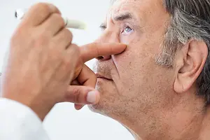 An elderly man uses a white dropper to administer eye drops into his eye.