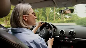 An older woman is driving a car with her eyes on the road and her hands on the steering wheel. She is wearing glasses and a blue shirt. She is probably on a road trip or going somewhere. She is looking at the side mirror, which reflects her face. The side mirror is attached to the car's window. There are trees and a road with a white line in the distance.