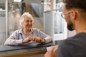 An elderly woman is smiling and talking to a man in glasses and a black shirt in an office building.