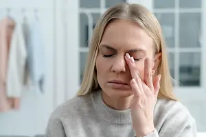 A woman with blonde hair and closed eyes is sitting on a couch in a room with a window in the background, holding her head with her right hand and looking to the left.