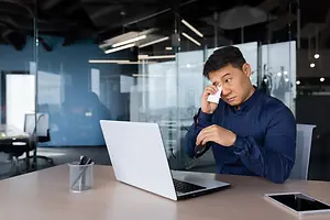 A man sitting at a desk in an office with a laptop and a tablet, holding a tissue to his nose.