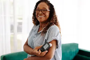 A smiling African American woman in glasses and a striped shirt is holding a glucose meter in her left hand, with a green couch and white curtains in the background.