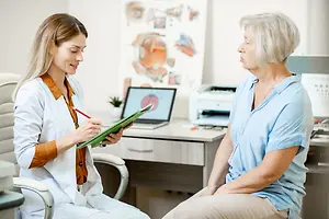 An older woman sitting on a chair while speaking to a female doctor who is taking notes on a clipboard