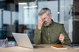 An older man is sitting at a desk, holding his head with his left hand, and seems to be looking at his laptop with a blurry view of the glass wall behind him.