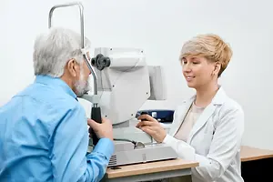 An old man is checking his eyes using a machine while a female doctor is smiling and holding a pen near him.