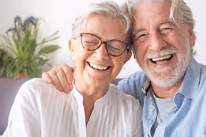 An older couple, both wearing glasses, smiling and posing for a picture in a living room.