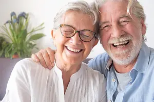 An older couple, both wearing glasses, smiling and posing for a picture in a living room.