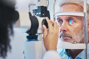 An older man wearing a blue shirt is having his eyes checked by a doctor using an ophthalmoscope.