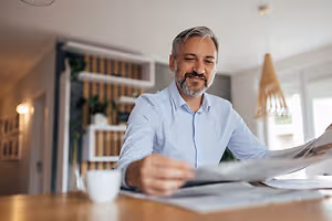 A man in a blue shirt is sitting at a table reading a newspaper and smiling