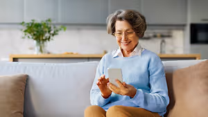 An older woman is sitting on a couch, smiling while holding a white tablet, wearing glasses and a blue sweater. Behind her is a kitchen with a plant on a wooden shelf, a microwave on the right, and a sink with a faucet.