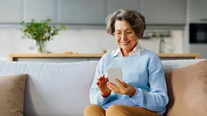 An older woman is sitting on a couch, smiling while holding a white tablet, wearing glasses and a blue sweater. Behind her is a kitchen with a plant on a wooden shelf, a microwave on the right, and a sink with a faucet.