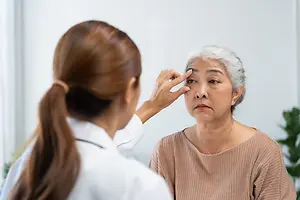 An elderly woman with gray hair is having her eye examined by a young woman doctor in a white coat.
