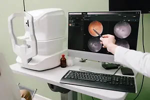 A doctor in a white coat using an ophthalmoscope to examine a patient's eye while looking at the monitor
