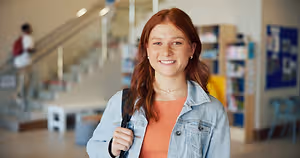 Portrait of a young woman wearing a denim jacket and carrying a backpack, standing in a library with a staircase and bookshelves in the background