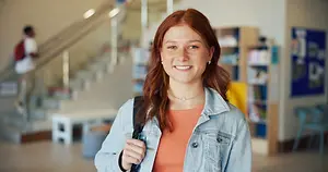 Portrait of a young woman wearing a denim jacket and carrying a backpack, standing in a library with a staircase and bookshelves in the background