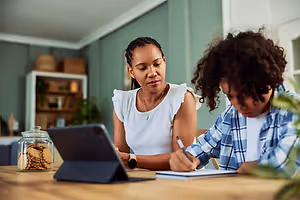 A woman and a child are sitting at a table with a laptop and a notebook.