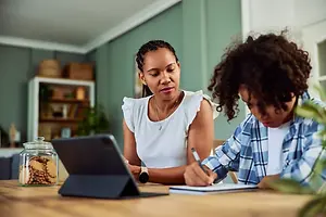 A woman and a child are sitting at a table with a laptop and a notebook.