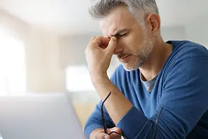 A man is sitting in front of a laptop and rubbing his forehead with his right hand. He has glasses on his left hand and is looking at something on his laptop. He is wearing a blue long-sleeve shirt with a white inner shirt. Behind him is a blurry view of a window and a white wall.