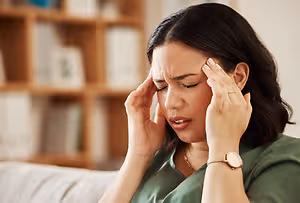 Woman with black hair sitting on a couch and looking distressed while holding her head