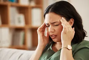 Woman with black hair sitting on a couch and looking distressed while holding her head