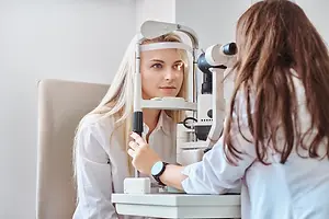 A woman wearing a white shirt is having her eyes examined by a female doctor in a white lab coat using an ophthalmoscope.